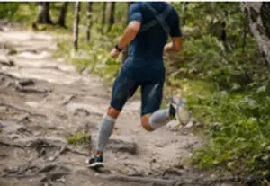 A person in mid-stride, running on a rocky trail in a natural setting. 3D printed insoles and compression stockings are worn for performance enhancement.